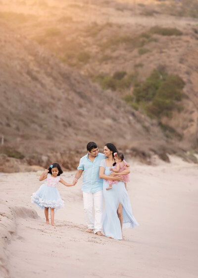 Family of mom, dad and two daughters walking along the beach in Malibu wearing pastel blue and pink colors. - Los Angeles Family Photography