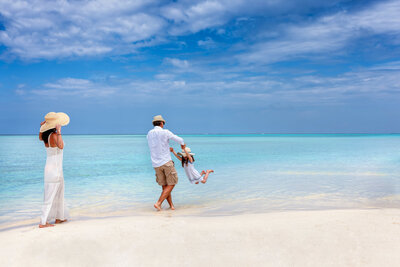 A family enjoys the beach; a woman in a white dress and hat stands near the shore, while a man swings a child by the hands. The sea is a vibrant blue, and the sky is partly cloudy, creating a serene and joyful atmosphere.
