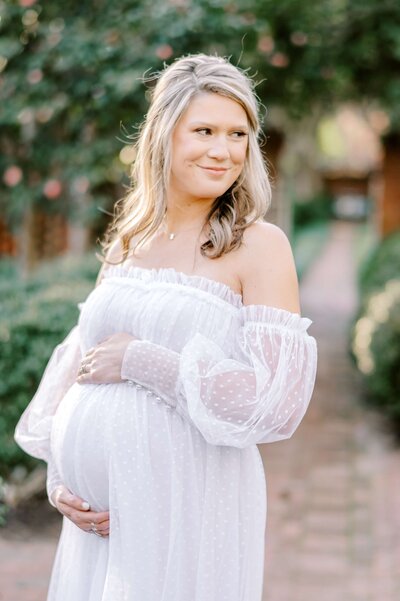 A pregnant woman in a romantic white tulle gown cradles her bump while smiling over to the side inside of Wing Haven Gardens + Estate in Charlotte, NC