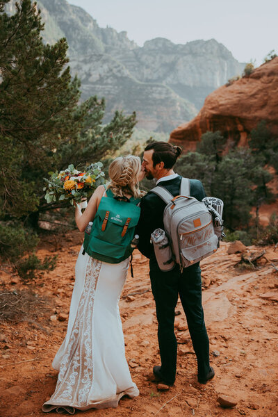 man and woman standing together kissing with backpacks on and wedding attire