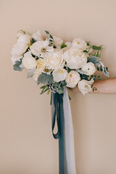A white and green bouquet held out against a cream colored wall with dark blue ribbon.