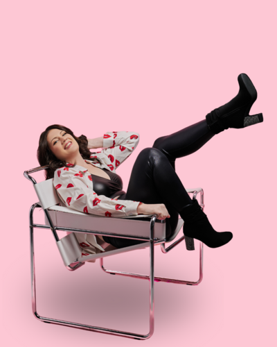 Emily is sitting in a low white chair leaning back with her legs in the air, showing off her black boots with sparkly heels and wearing a red lip pattern shirt on a light pink background