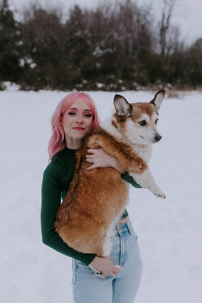 A girl with pink hair sits on the ground holding her corgi breed dog in her lap