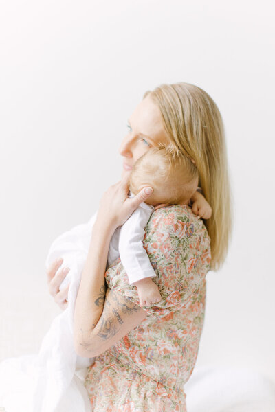 A blonde mother holding her newborn baby boy over her shoulder while staring out the window during photo session with Boston newborn photographer Corinne Isabelle
