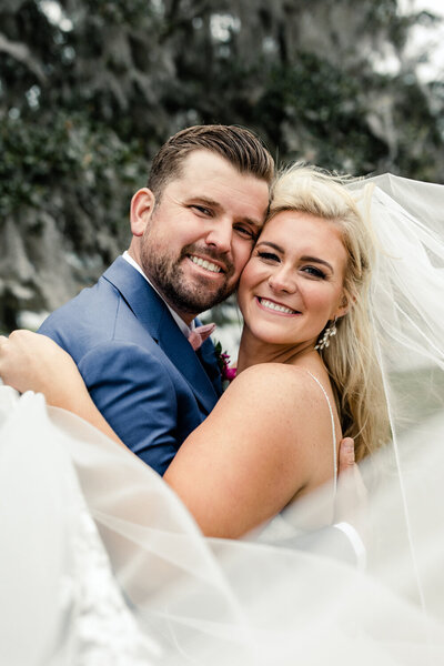 Bride with blonde hair in white gown and veil hugging groom in blue suit with brown hair by Charleston wedding photographer, Stephanie Bailey Photography
