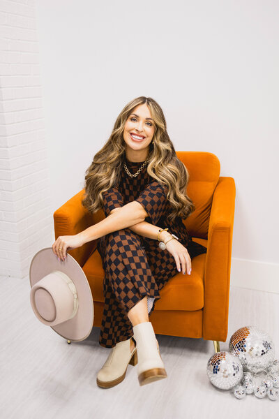 woman holding a wide brimmed hat while sitting in an orange chair and smiling