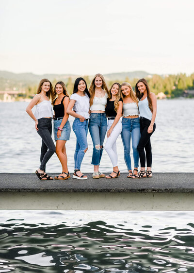 A group of high school  senior girls stand with arms. around each other on a riverside dock