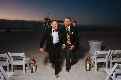 two grooms walking down wedding aisle while smiling