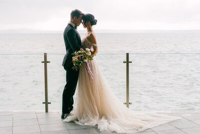Bride and groom stand facing together intimately on deck overlooking the ocean