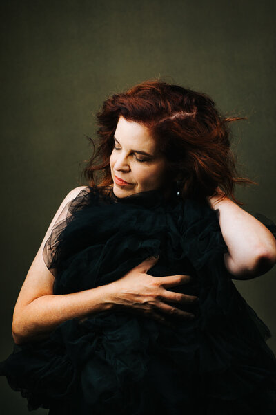 Redheaded middle-aged woman holds a bundle of black tulle over her torso. She's smiling and looking down, and one hand reaches back to touch her neck.