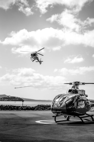 A black and white photo of a helicopter on the ground and another in the air flying back to base to land captured by Auckland Commercial Photographer - Zanthe Vorsatz Photography