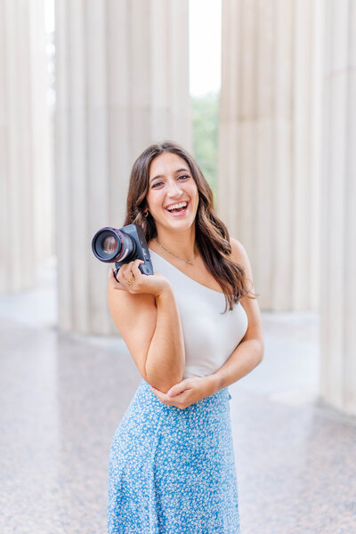 Nashville wedding photographer, Brooke elliott posing with her camera for a branding photo wearing a white tank top and blue floral skirt under the Nashville War Memorial