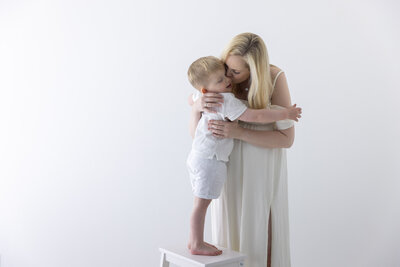 Studiofoto av en gutt som står på en hvit krakk og gir moren sin en klem. Mor iført hvit kjole, kysser sønnen på kinnet hans.