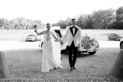 Newlyweds couple arriving at the location for the reception. They are smiling at the camera walking. He is swearing a tuxed white and black, she is wearing a luxury wedding dress with long sleeves covered by lace. Behind them an historic car. 