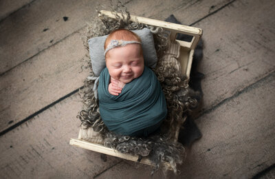 Newborn girl smiling while wrapped in teal blue wrap laying on grey curly rug on white baby bed