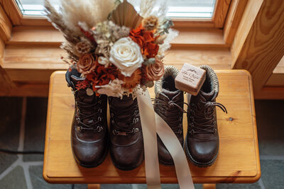 Two pairs of rugged brown hiking boots, one with cozy fur lining, are neatly placed on a wooden bench, symbolizing the adventurous spirit of the couple whose wedding they're prepared for. A beautiful bridal bouquet, filled with an array of flowers including deep orange and white roses, with pampas grass, is propped inside one of the boots, adding a touch of wild elegance to the ensemble. A wooden ring box, engraved with the names "Adam & Jess" and a mountain range, sits atop the other boot, signifying the couple's shared love for the outdoors. The warm indoor lighting and wooden interior hint at a cozy, intimate setting, possibly a cabin, perfect for a rustic wedding theme.