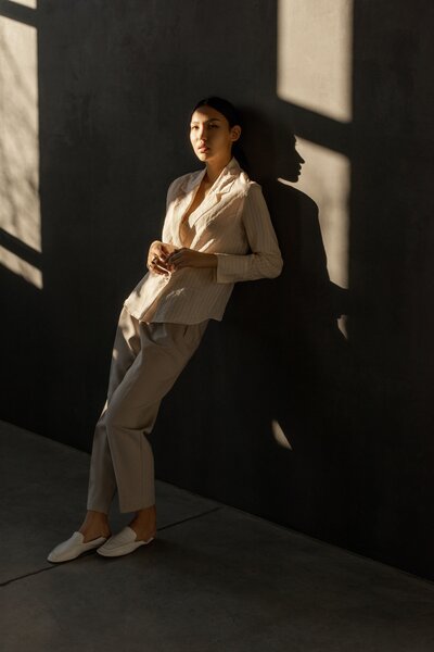 A woman in a light tailored suit with loafers and hair tied back leaning against a wall  with dark shadows and light through a window.