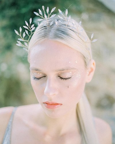 ethereal image of white woman with blond hair, closing her eyes with gold flecks on eyelids, cheekbones and temples. She is wearing a gold laurel leaf crown in her hair and a spaghetti strap silvery white gown.