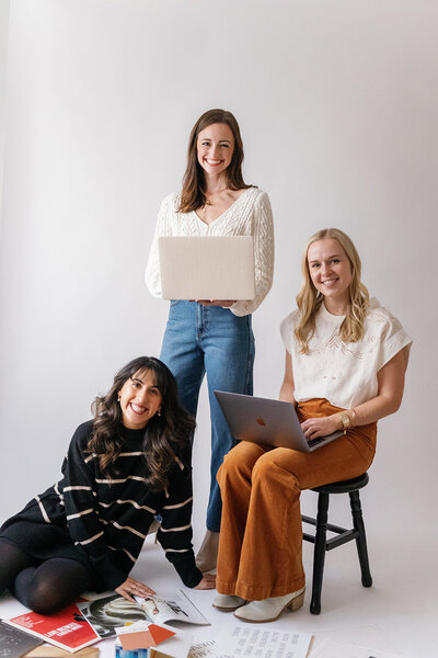 One woman with a laptop standing above another woman with a laptop in a chair and another woman sitting on the floor
