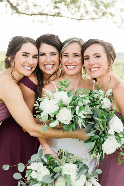 Bridesmaids hugging bride and smiling at camera while holding white and green bouquets