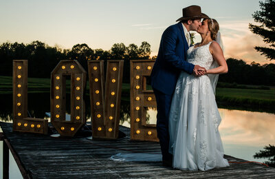 Bride and groom kissing next to lighted LOVE sign at sunset during their wedding reception in Waterford, PA