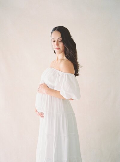A woman wearing a white dress and holding her pregnant belly during portraits in NJ