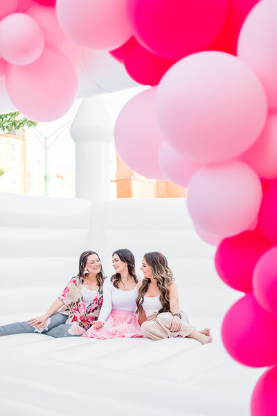three brunette women sitting in white bounce house with pink balloon garland