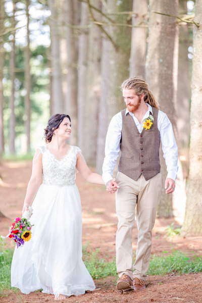 Bride and groom hold hands and smile while walking amidst trees holding bouquet