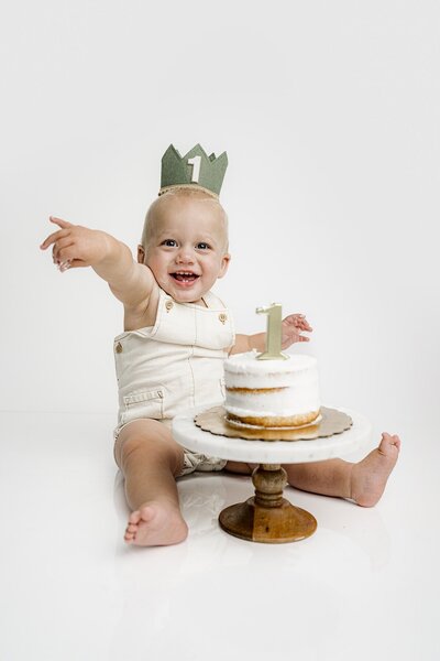 little boy sits wearing a crown playing in his first birthday cake
