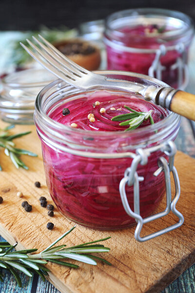 A jar of pickled red cabbage sits next to a fork and fresh rosemary sprigs on a wooden surface.