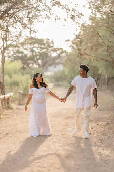 Expecting couple in all white walks hand in hand down a path smiling at each other.