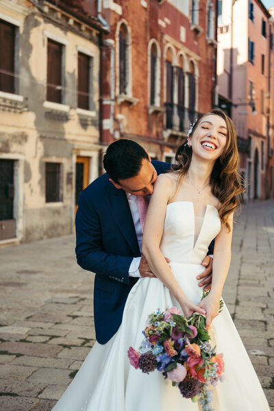 Newlyweds in Venice captured in a candid moment. Groom kissing bride's shoulder as she holds a colorful bouquet. Quaint Venetian alley in the background creates a charming atmosphere. A picture-perfect memory of a dreamy Venetian wedding.