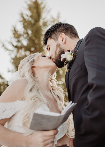 blonde bride in an off the shoulder wedding  dress and groom in a black suit kiss while the bride is holding her vows in a little book, captured by Jackson Hole wedding photographer