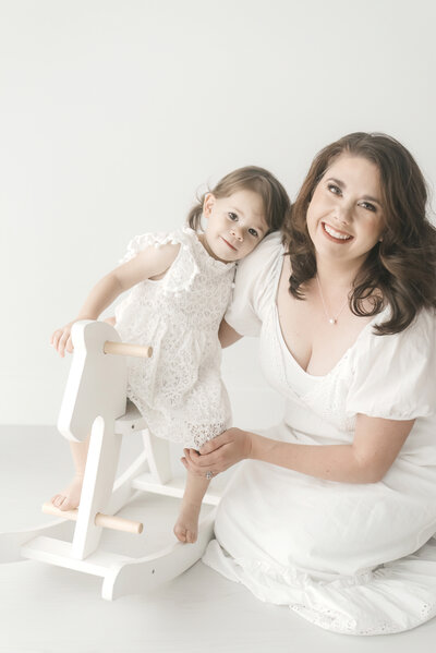 Mother and child posing next to white rocking horse in white dresses