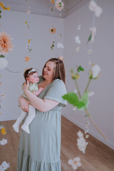 Mom wearing green dress holding baby daughter wearing a matching dress surrounded by flowers in a photography studio
