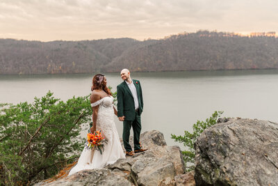 A wedding couple standing on a rock overlooking a lake.