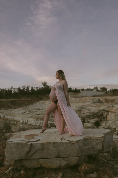 Expectant mother standing barefoot on a rocky ledge during a maternity photoshoot, holding her belly as her pink gown flows in the wind at sunset.