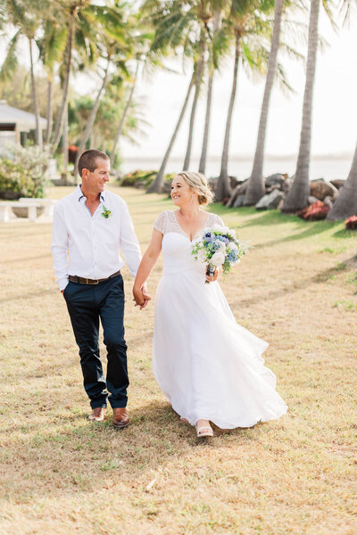 Bride and groom holding hands as they walk under palm trees of their beach wedding.