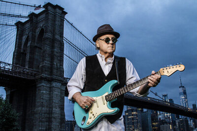 Musician photo Robert Allen standing in front of Brooklyn Bridge wearing sunglasses and black hat while holding blue electric guitar at sunrise