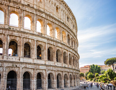 The image shows the Colosseum in Rome, Italy, lit by daylight. The ancient amphitheater's stone arches and tiers are visible, with people walking nearby and green trees in the background under a clear blue sky.