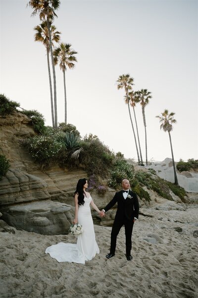 Elegant Southern California beach wedding in Laguna with bride and groom holding hands on the sand, surrounded by coastal cliffs and palm trees
