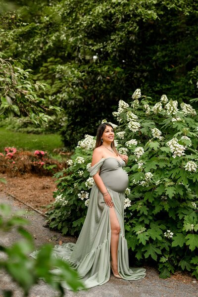 Charlotte Area mother stands in front of flowers at Glencairn Gardens wearing a green maternity two-piece dress