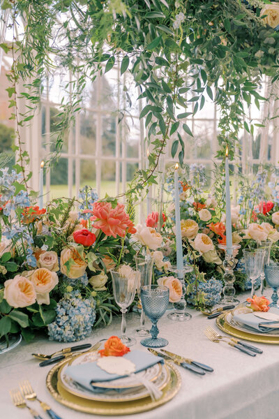 luxury wedding table design with abundant blue and coral flowers and layered gold and white plates topped with a blue folded napkin