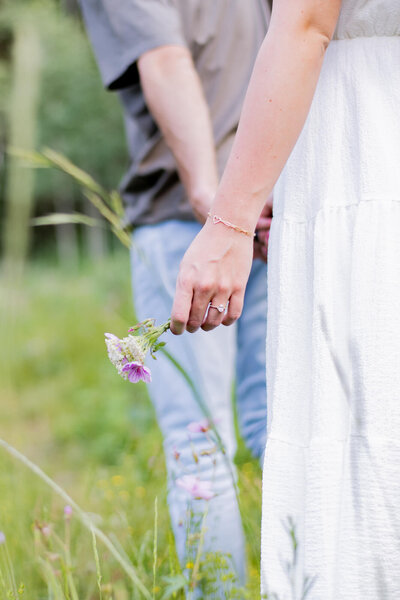 Couple holding hands and walking away, but the picture is focused on the girl's ring hand that is holding a little group of flowers