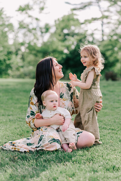 A mother wearing a floral dress sitting in the grass with her baby son and her toddler daughter