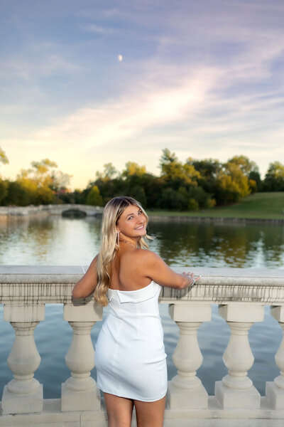 a blonde haired girl in a tight white dress  standing by a cement railing  next to a lake at  The Grand Basin in Forest Park at sunset.