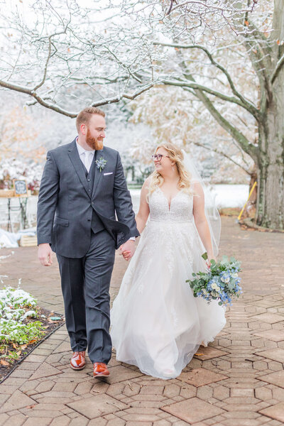 A bride and groom walk together holding hands. She is holding her bouquet and looking up and smiling at him as he smiles at her. It has snowed and they are walking on a brick path