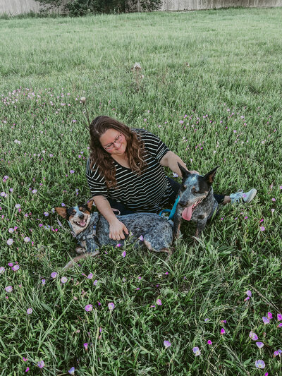 A brunette plus size woman in Alvin Texas sitting in a field of wildflowers with two australian cattle dogs