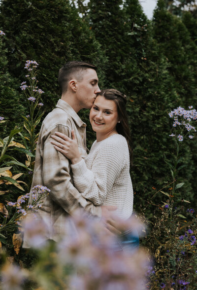 Knoxville photographer captures outdoor engagement photo with couple embracing in a garden with lavender flowers surrounding them as the man kisses the womans head and she smiles at the camera