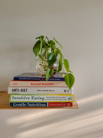 Intuitive Eating and Anti-Diet books sit stacked on a shelf with a green plant sitting on top
